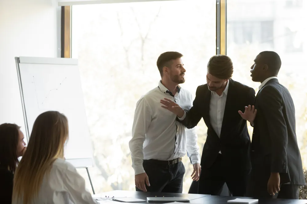 Two male coworkers arguing during a tense meeting while a third man tries to separate them, with others watching nearby.