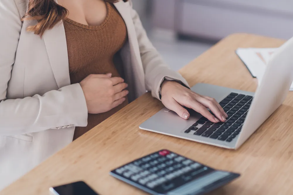 Pregnant woman working on a laptop at a desk, with one hand on her belly.