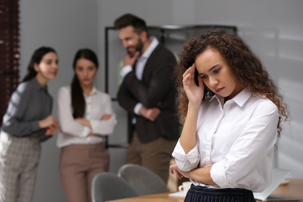 Upset woman standing apart from a group of coworkers who appear to be gossiping and watching her in the background.