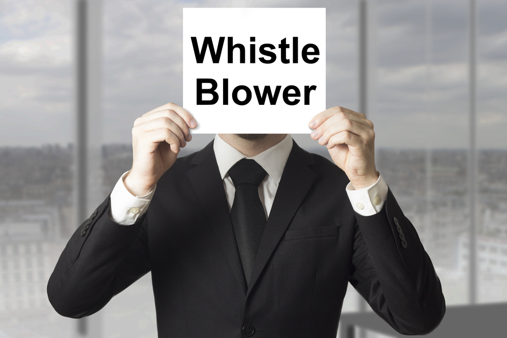 Man in a suit holding a sign in front of his face that says "Whistle Blower."