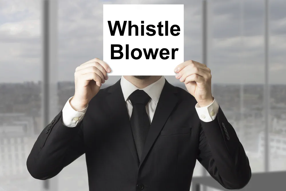 Man in a suit holding a sign in front of his face that says "Whistle Blower."
