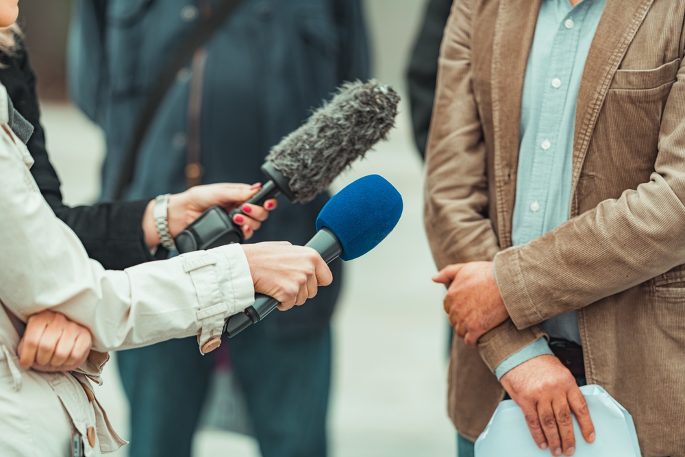 Person speaking to reporters holding microphones during an interview.