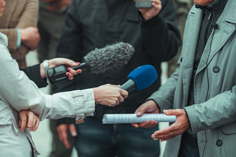 A group of journalists holding out microphones during a press interview with a man wearing a grey coat.