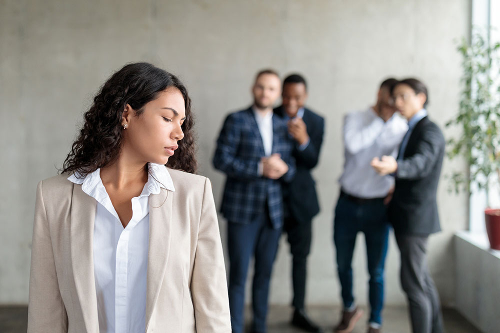Female employee looking isolated while male coworkers whisper behind her, illustrating subtle workplace discrimination in the office
